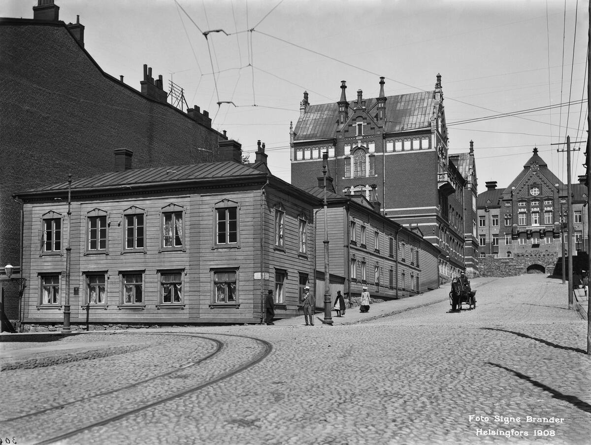 Nikolainkatu (= Snellmaninkatu ) 16, 18. Etualalla matala puutalo, sen takana uusia kivitaloja. Mukulakivikadulla ajaa mies hevosvaunuilla.