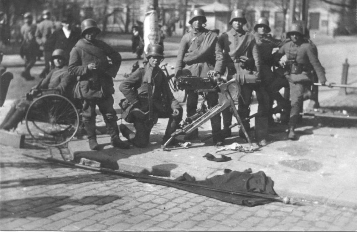 Ryhmä saksalaisia sotilaita kadulla. Heidän edessään maassa makaa punakaartin lippu.