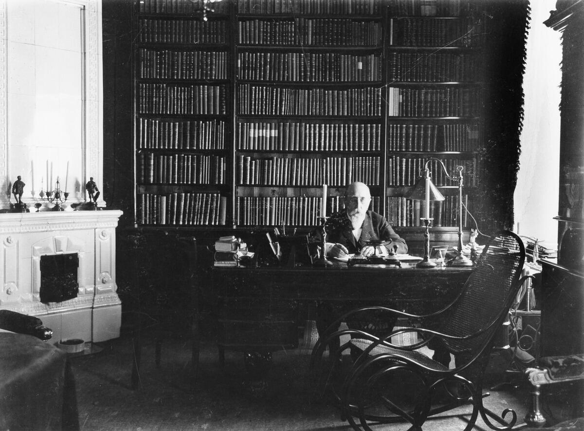 En man sitter bakom sitt skrivbord och har fotograferats framifrån. Bakom honom finns en bokhylla som täcker hela väggen och till vänster en vit kakelugn.