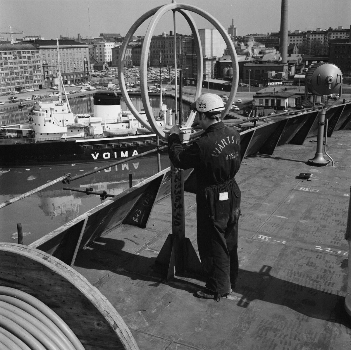 På väderdäcket till ett kryssningsfartyg under konstruktion står en arbetare vars overall har texten Wärtsilä på ryggen. I bakgrunden syns isbrytaren Voima, och bakom den Sandvikstorget, Sinebrychoffs bryggeri och höghus.