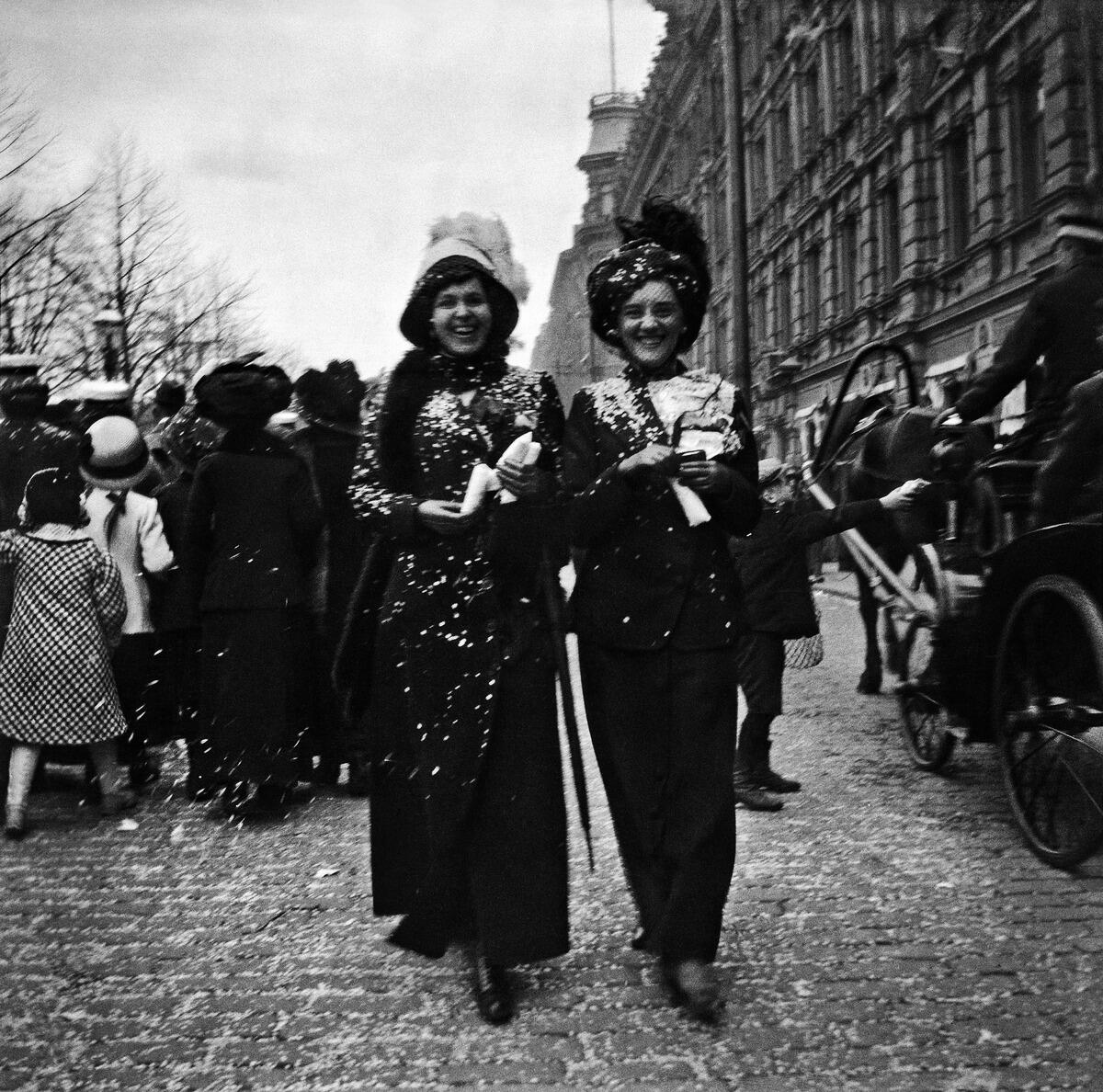 Två storleende kvinnor promenerar längs Esplanaden. I bakgrunden syns massor av människor. Damerna har konfetti på sig, och konfetti finns också på gatstenarna.