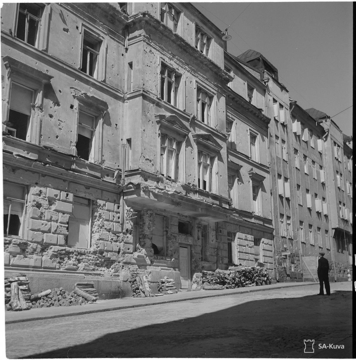 I förgrunden syns byggnadens illa tilltygade fasad. På gatan står en mänska i skuggan av byggnaden och betraktar ödeläggelsen. 