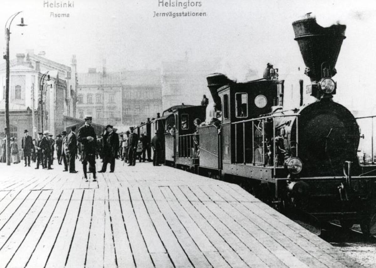 Ånglok med passagerarvagnar vid Helsingfors gamla järnvägsstation som byggdes 1862 och revs 1918. Människor står på perrongen.