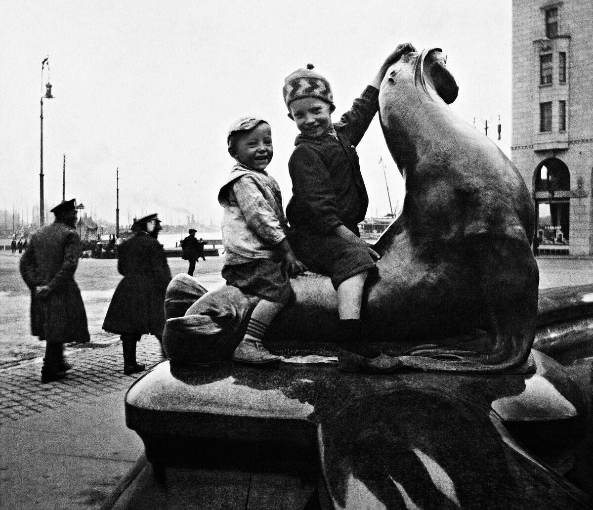 Kaksi lasta ratsastaa hylkeen selässä Havis Amanda -patsaalla Kauppatorilla. Taustalla näkyy ohikulkijoita ja meri.