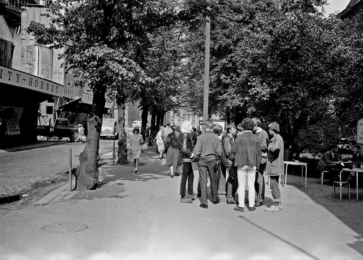 Ryhmä nuoria seisoo puistikossa, joka sijaitsi Ateneumin kulmalla. Vasemmalla on työmaa-aita, jonka takana rakennetaan Makkarataloa.