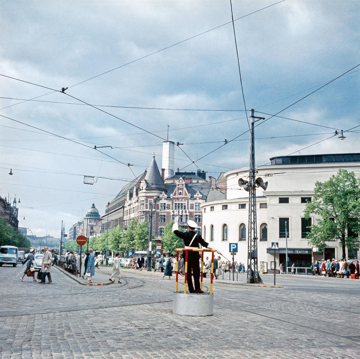 Fotgängare korsar gatan vid korsningen av Bulevarden och Mannerheimvägen. Vid Skillnaden står en trafikpolis på en plattform och vinkar med händerna.