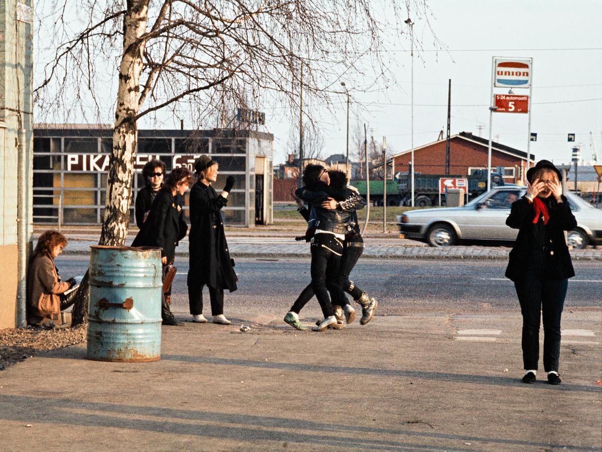 Henkilöt halailevat toisiaan. Nuoriso on pukeutunut punk-henkisesti. Taustalla toisella puolella tietä on autopesula ja Union-huoltoasema. Vasemmalla näkyy ohut kaistale Lepakkoa. Lepakko eli entinen Lepakkoluola oli Helsingin Ruoholahdessa sijainnut varastorakennus, joka tuli kuuluisaksi toimiessaan vuosina 1967–1979 asunnottomien alkoholistien hätämajoitustilana ja sen jälkeen tärkeänä nuorisokulttuurin tapahtumapaikkana. Rakennus purettiin syksyllä 1999.