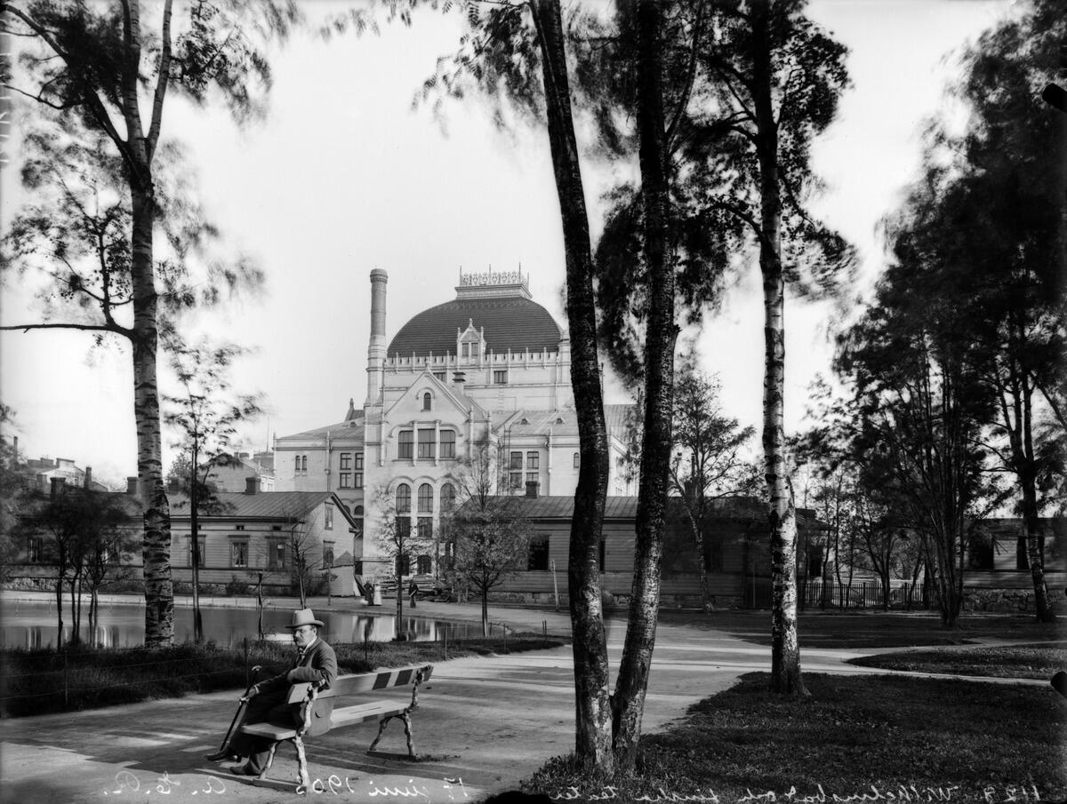 Kaisaniemen puisto, mies istuu penkillä. Vasemmalla joutsenlampi, taustalla Kansallisteatteri ja Villensaunan (Wilhelmsbad) kylpylän puurakennuksia.
