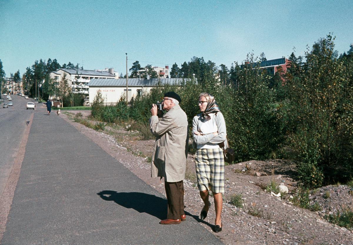 En man står sidledes hållande en kamera framför ögonen. Höger om honom går en kvinna med rutig kjol och kofta samt en huvudduk och solbrillor. I bakgrunden syns vita byggnader, i högra kanten buskage. 