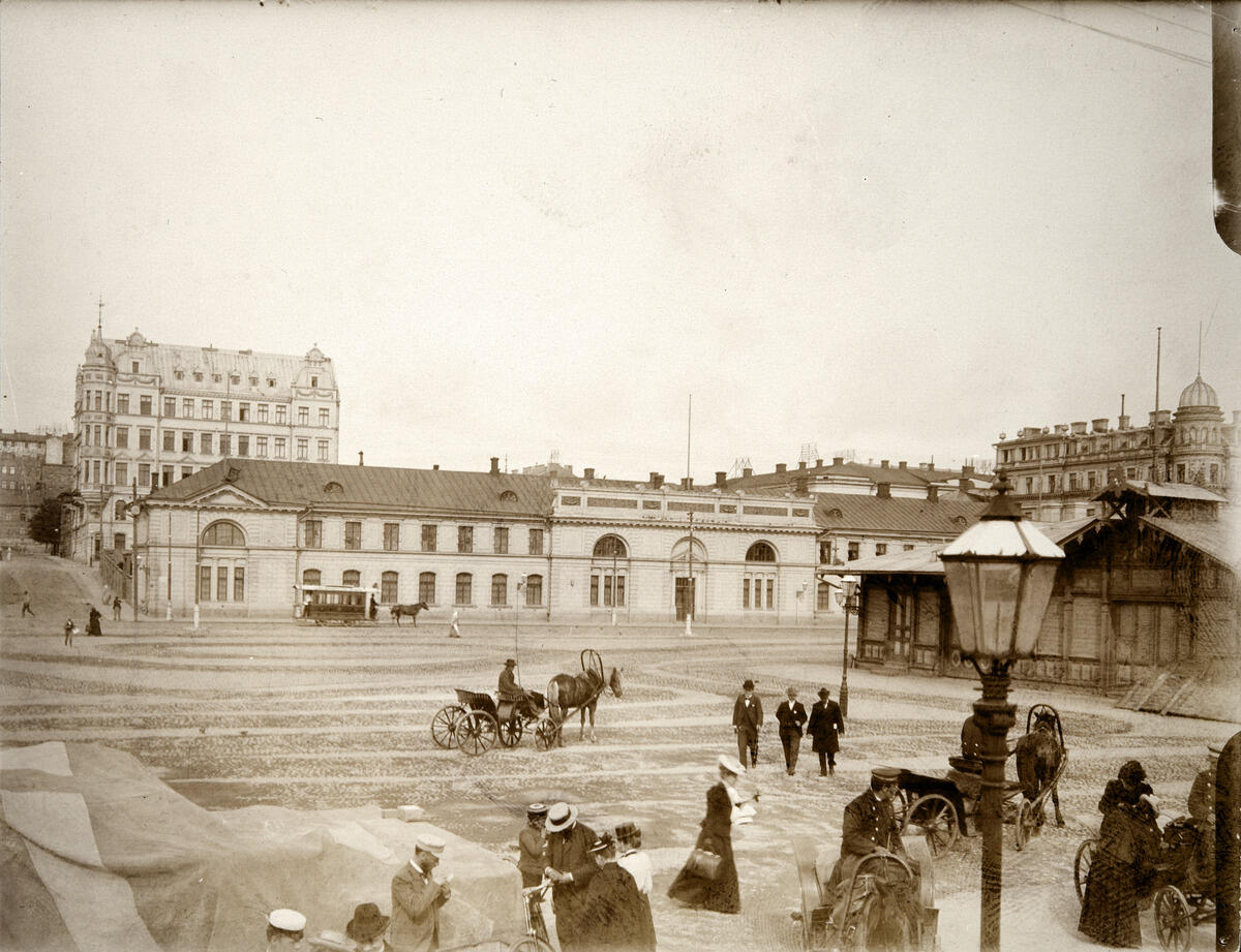 Hyrvagnar med sina hästar och herrskap vid Södra hamnen på Packhustorget. I bakgrunden det tidigare arméförrådet, från 1888 packhuset, Västra Hamngatan 10 (nuvarande Södra kajen 10). På gatan en hästdragen spårvagn. Bakom förrådet syns ett femvåningshus från 1960-talet vid hörnet av Unionsgatan 14 – Södra Magasinsgatan 3.