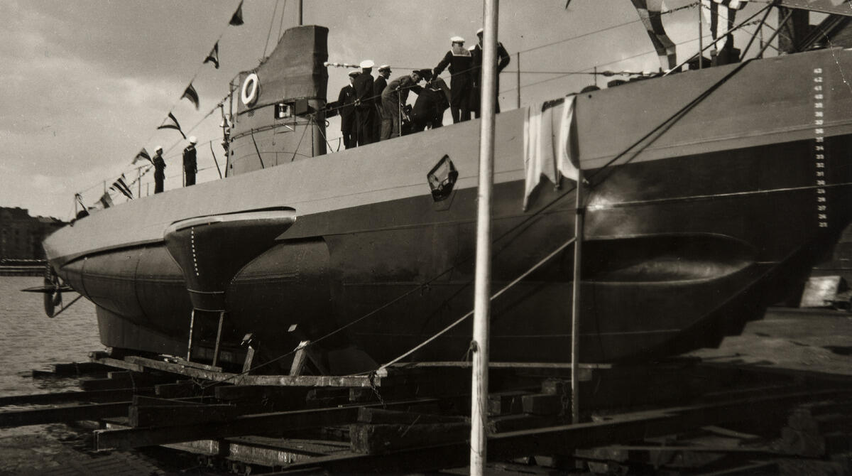 En ubåt fotograferad nerifrån. På däcket står marinsoldater i uniformer.