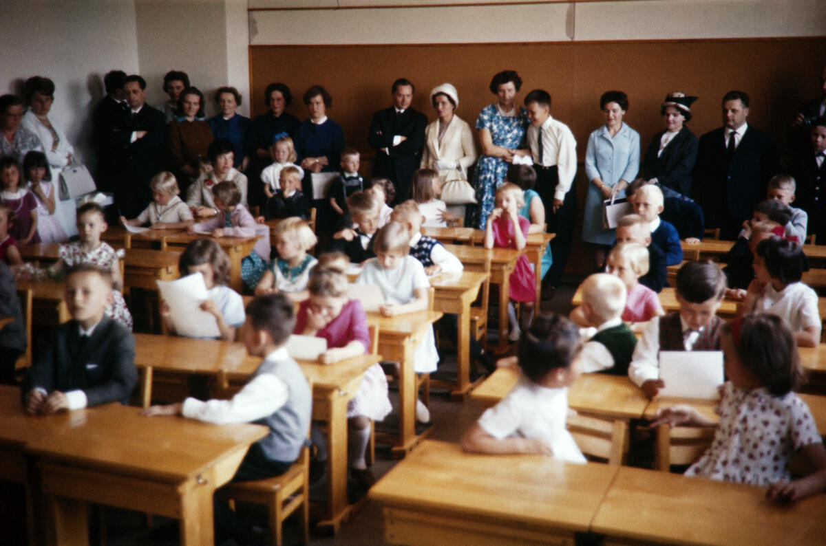 Vårfest i ett klassrum. Barnen sitter i pulpeterna och granskar sina betyg. I bakgrunden står föräldrar.