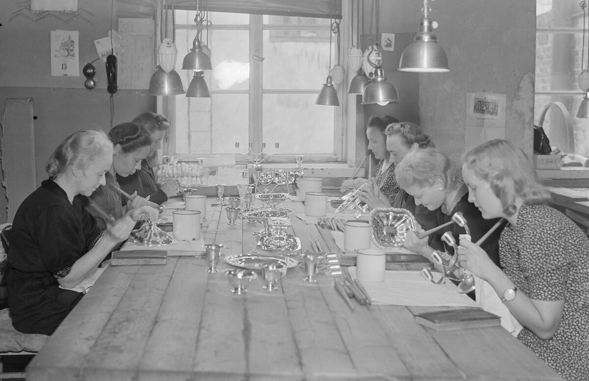 Seven women working on silverware around a large work table.