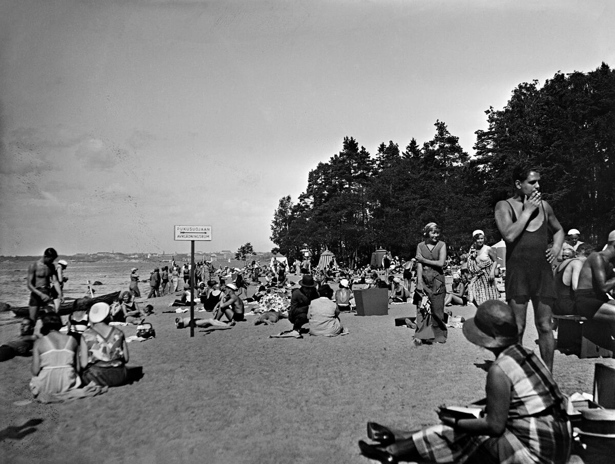 På stranden finns många människor. Vissa har badkläder, andra har klänning eller kostym och hatt.