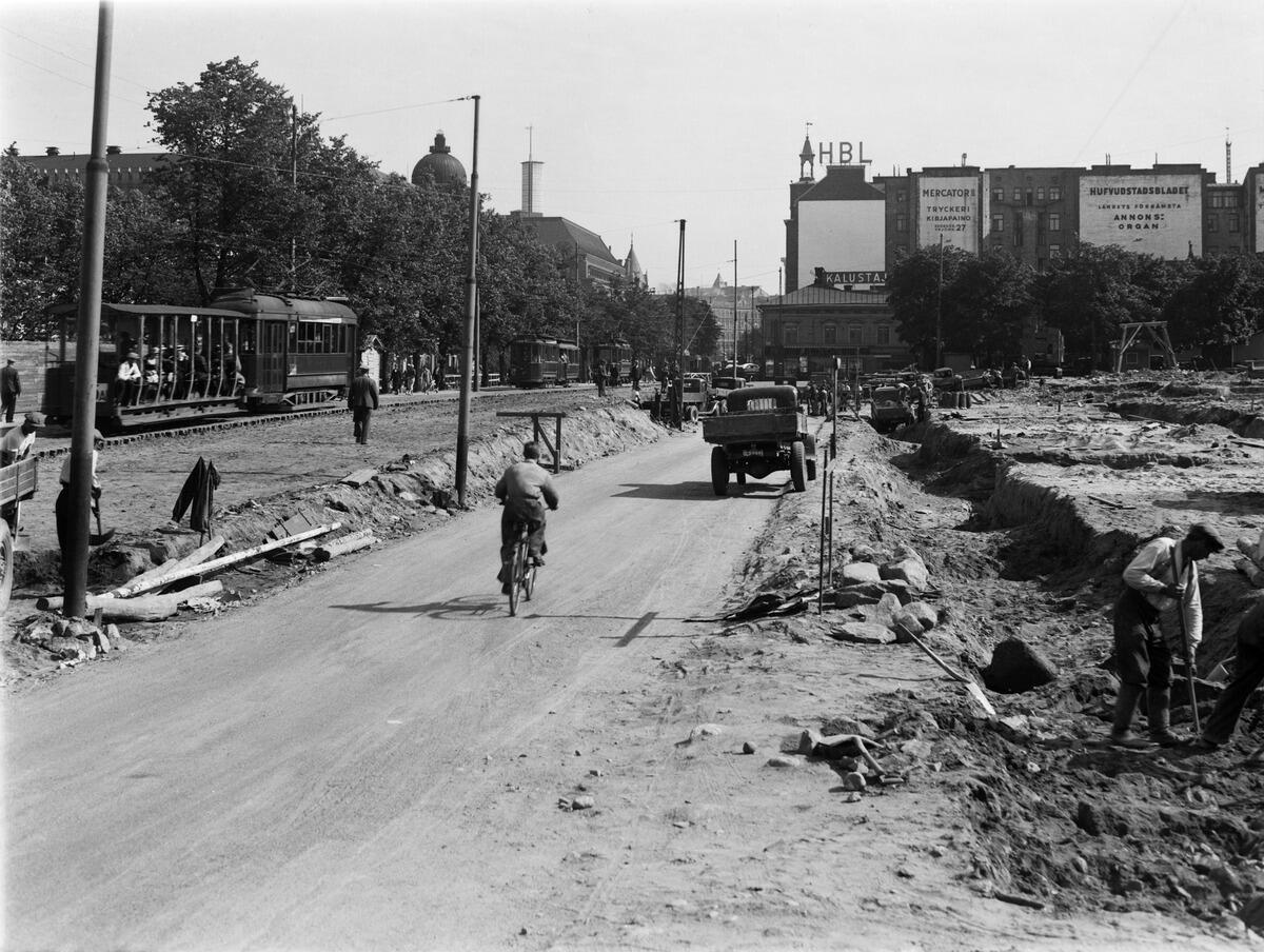 Ombyggnadsarbete på Henriksgatan (Mannerheimvägen). Till höger byggs Glaspalatset. Till vänster en spårvagn tillverkad av ASEA. Dess öppna bakvagn tillverkades av Ab Sandvikens Skeppsdocka och Mekaniska Verkstad.