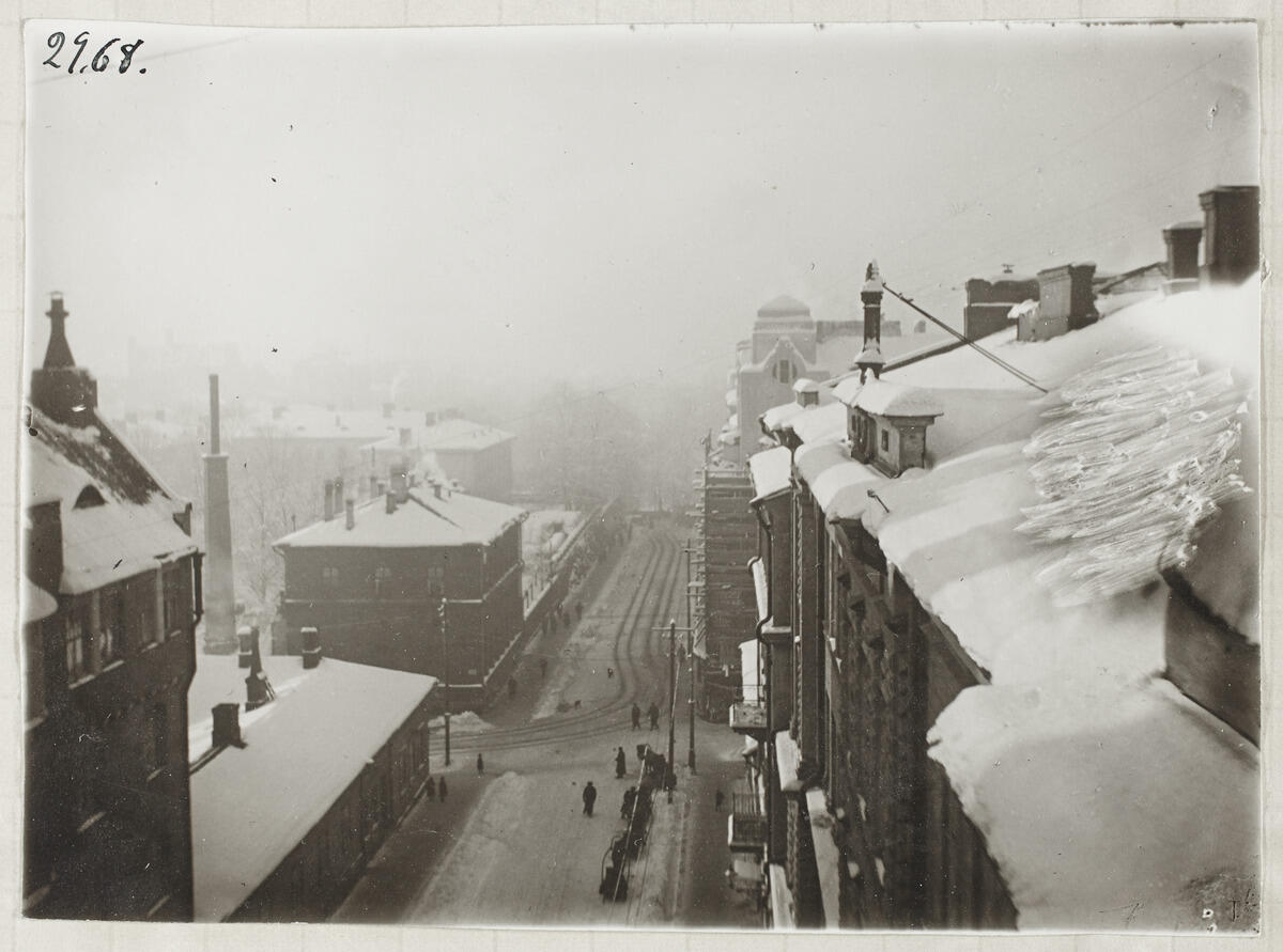 Korkealta ikkunasta otetussa kuvassa näkyy lumisia kattoja ja savupiippuja, alhaalla kävelee ihmisiä kadulla.
