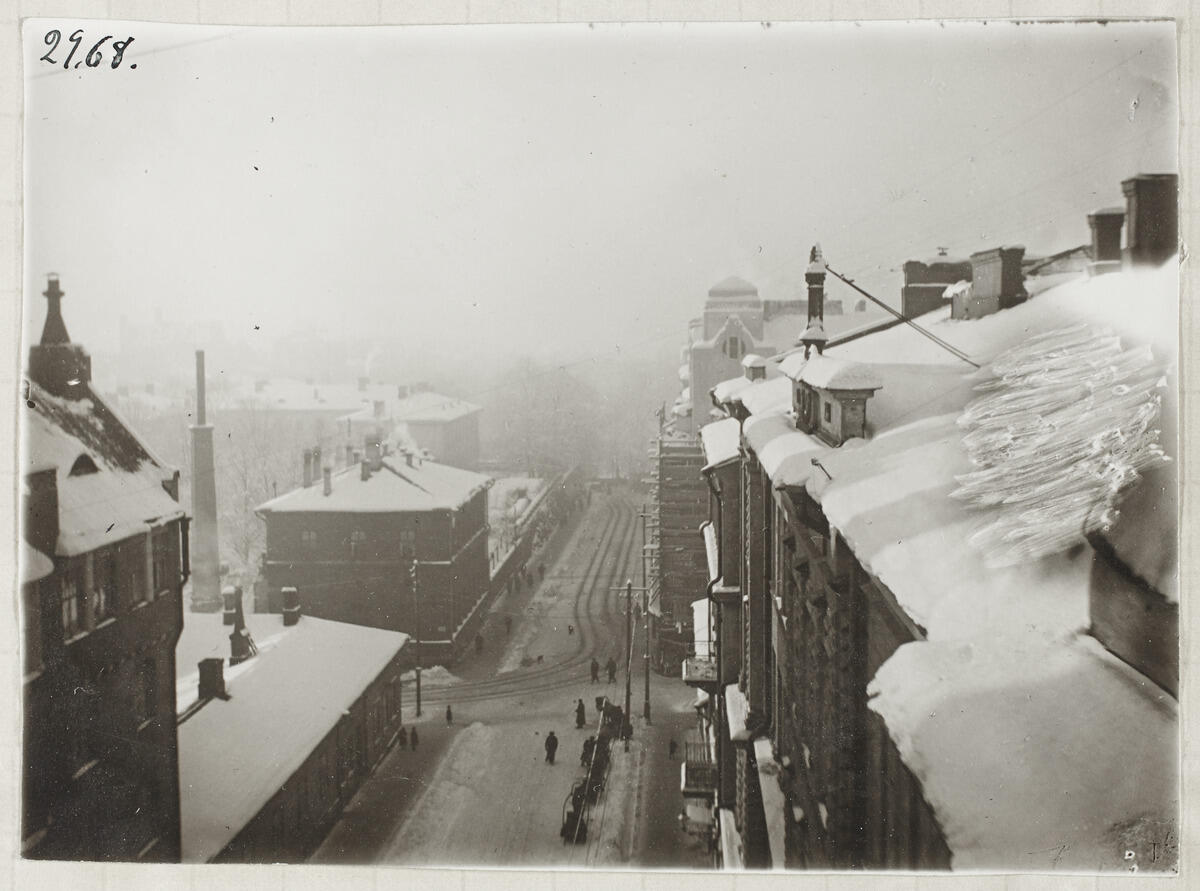 A photo taken from a window high up shows snow-covered roofs and chimneys, with people walking along the street below.