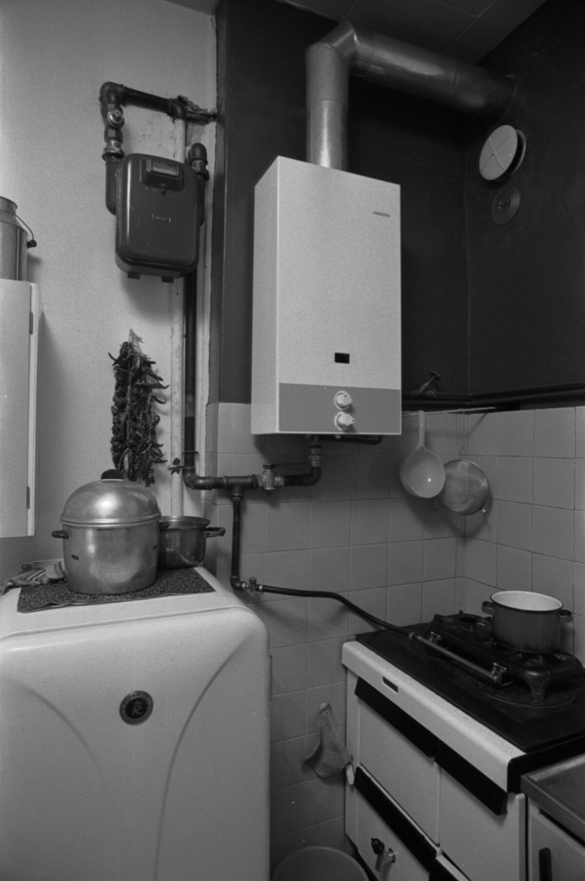 Keittiön nurkkaus, jossa kaasumittari, kaasukäyttöinen lämminvesivaraaja, kaasuliesi ja jääkaappi.