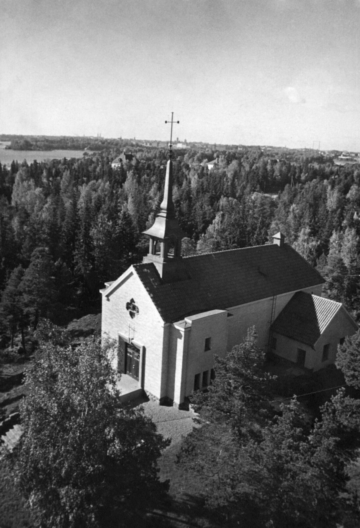 Yläviistosta kuvattu valkoinen kirkkorakennus metsäisessä ympäristössä.