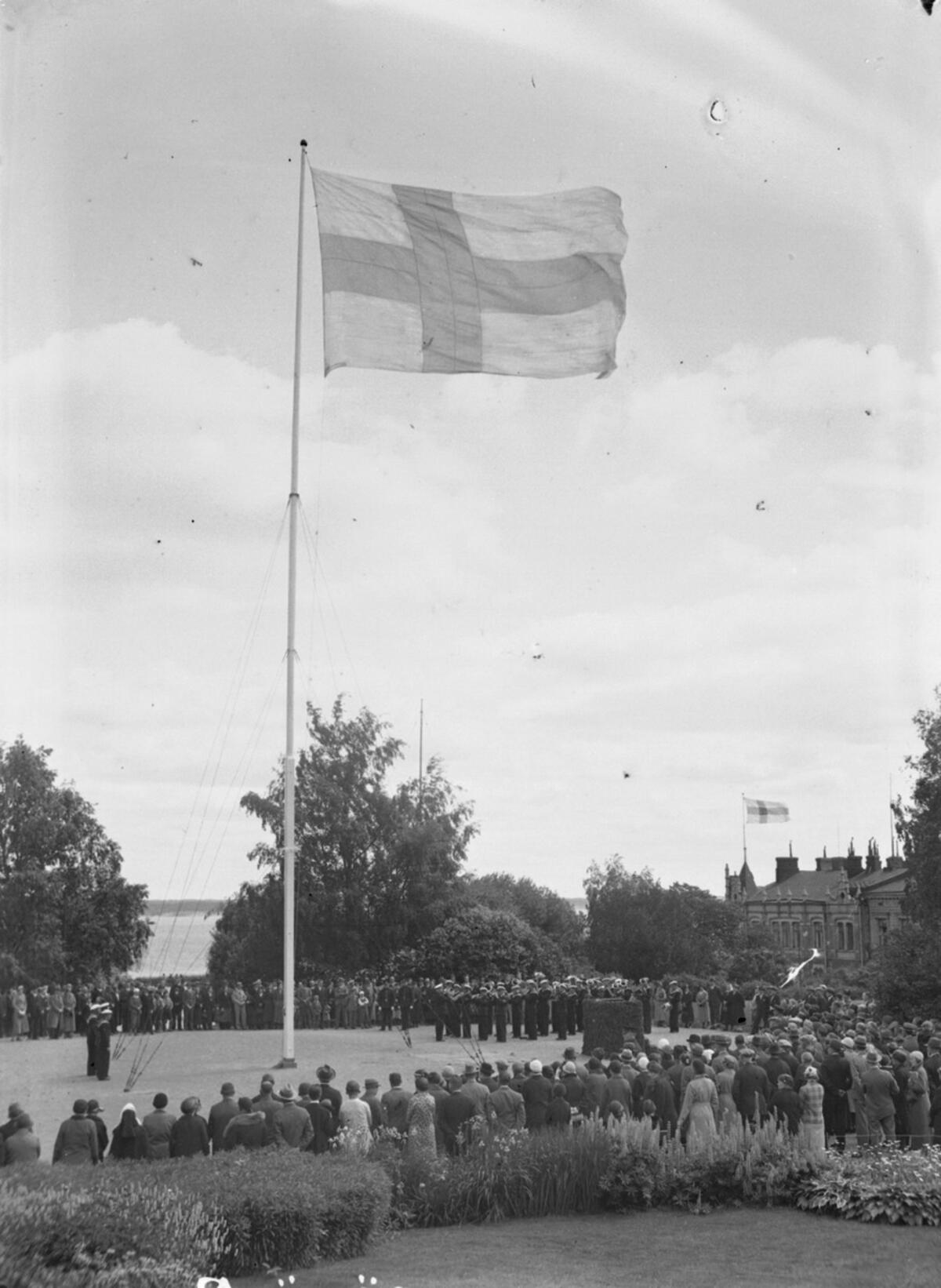Mustavalkoisen valokuvan keskellä  on lipputanko, jossa liehuu Suomen lippu. Ympärille on kokoontunut paljon ihmisiä.