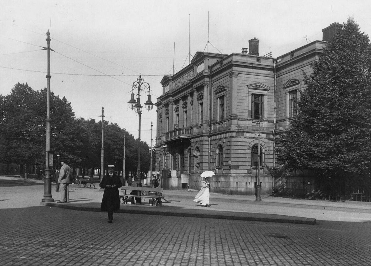 Vanhan ylioppilastalon edessä on koristeellinen kaasulyhty. Kadulla kävelee nainen, jolla on pitkä valkoinen mekko ja valkoinen päivänvarjo sekä mies, jolla on ylioppilaslakki.