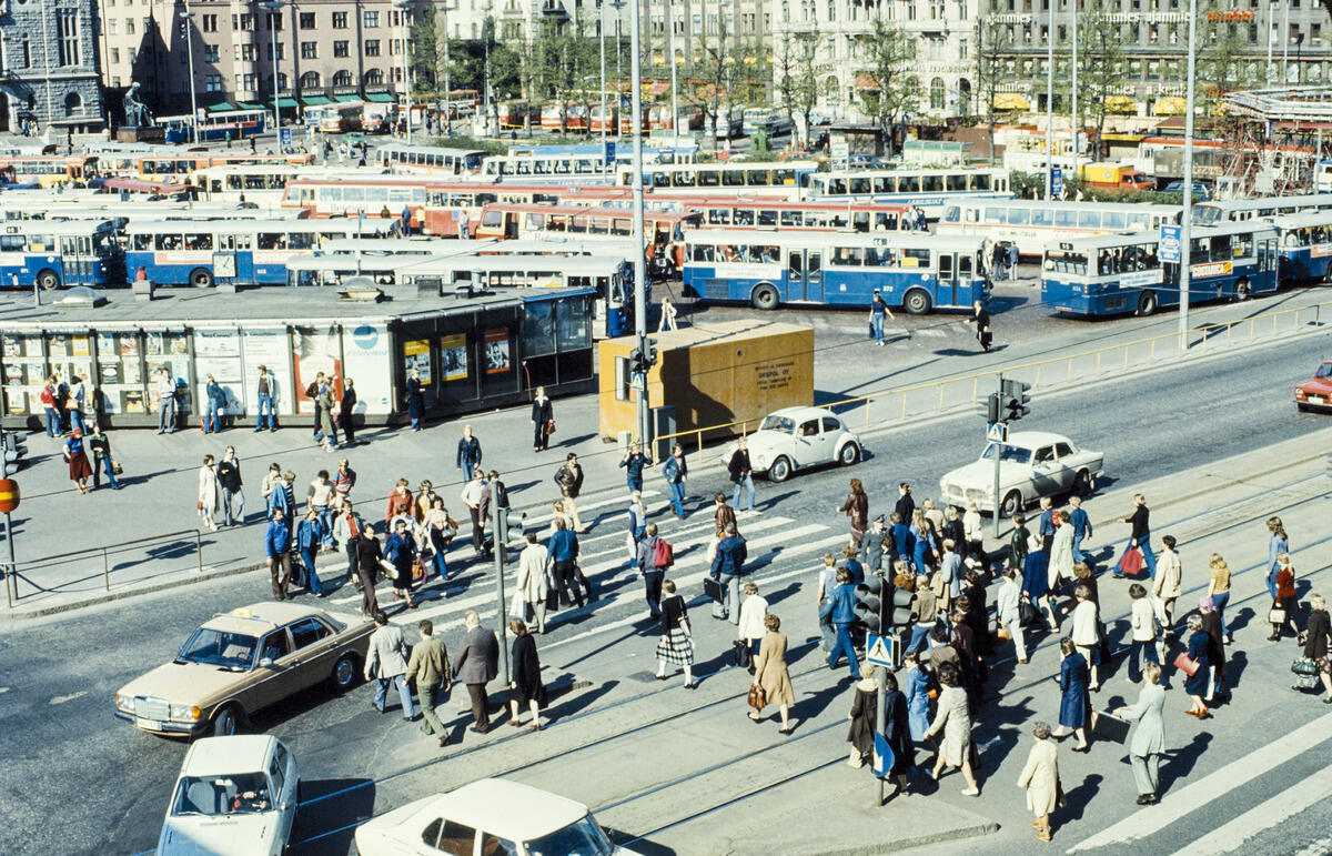 Jalankulkijoita ylittämässä suojatietä Kaisaniemenkadun alkupäässä, taustalla Rautatientorin bussipysäkkejä ja bussiliikennettä. Kuvan oikeassa ylänurkassa näkyvä putkirakennelma liittyy metrotunnelin louhintatöihin, joita tehtiin 1976–1977 ns. Kluuvin ruhjeen kohdalla. 