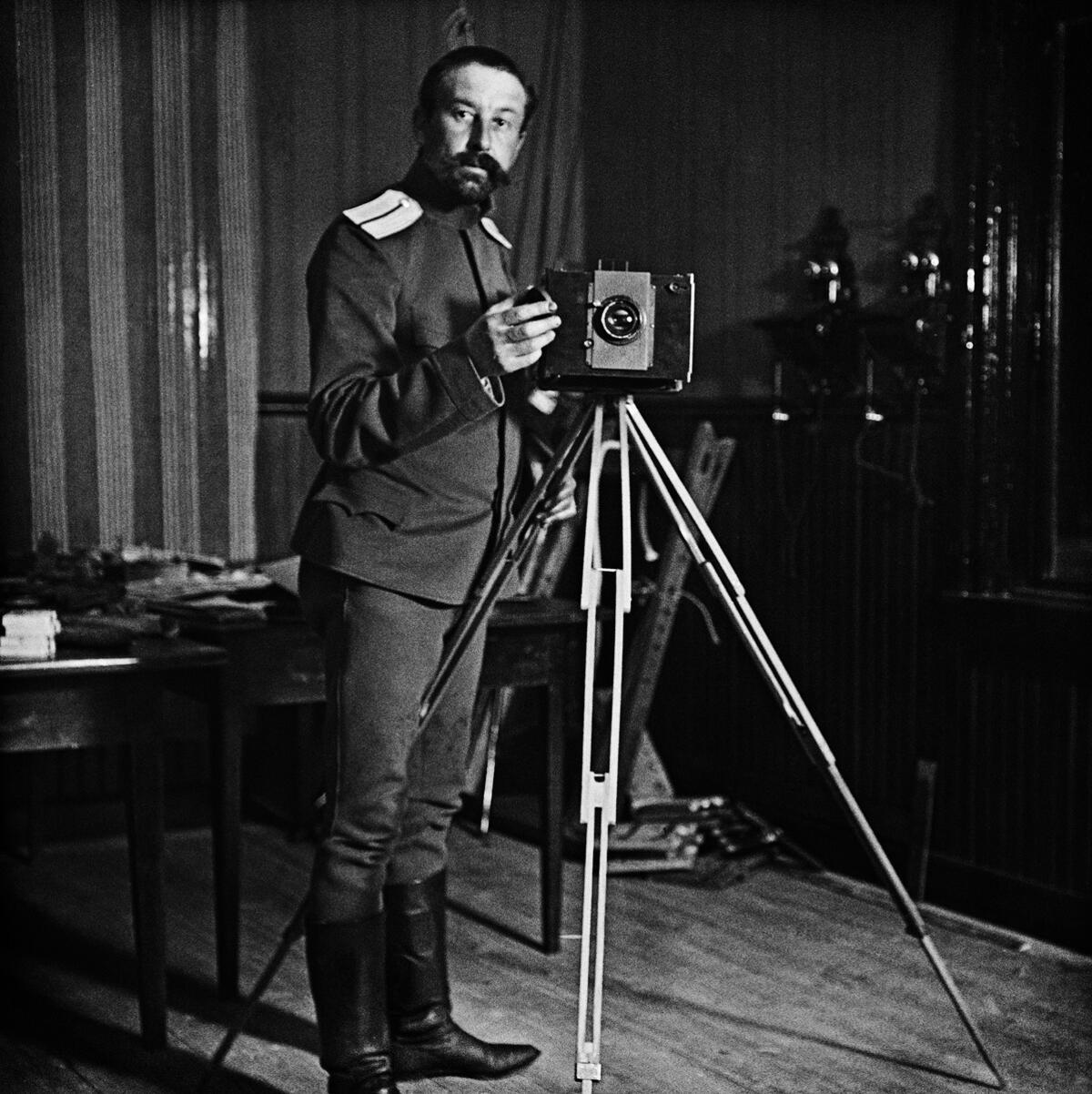 Univormupukuinen, parrakas ja viiksekäs mies seisoo jalustalla seisovan kameran takana. Taustalla näkyy seinä, johon on kiinnitetty kaksi puhelinta, sekä kaksi pientä pöytää, joilla on paperipinoja.