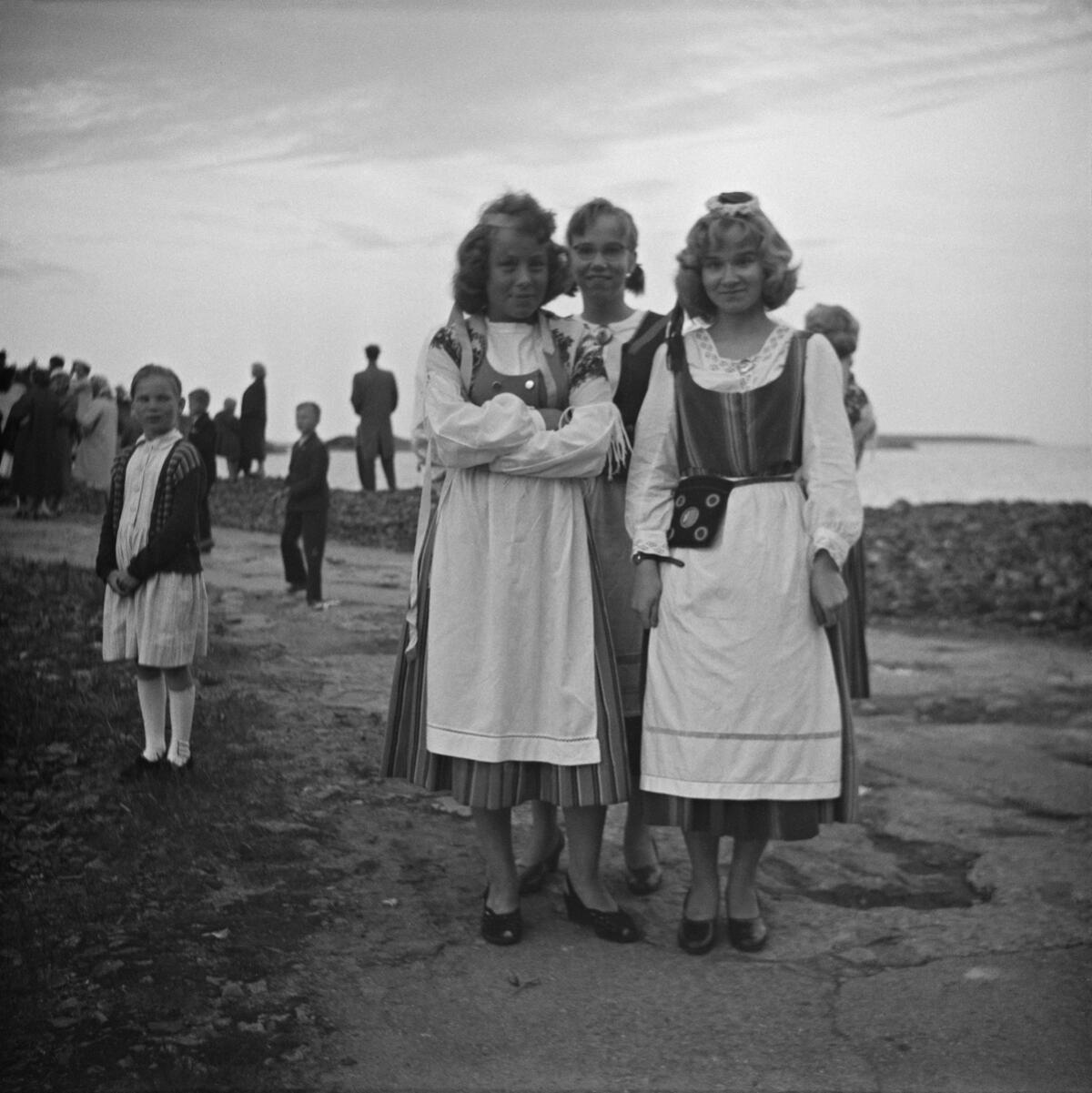 Two young women dressed in national costume pose in Vallisaari. A crowd watching the burning of the bonfire can be seen in the background.