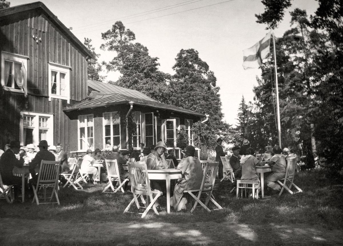 Ihmisiä istuu kahvilan ulkopuolilla pienten pöytien ääressä. Lipputangossa liehuu Suomen lippu.