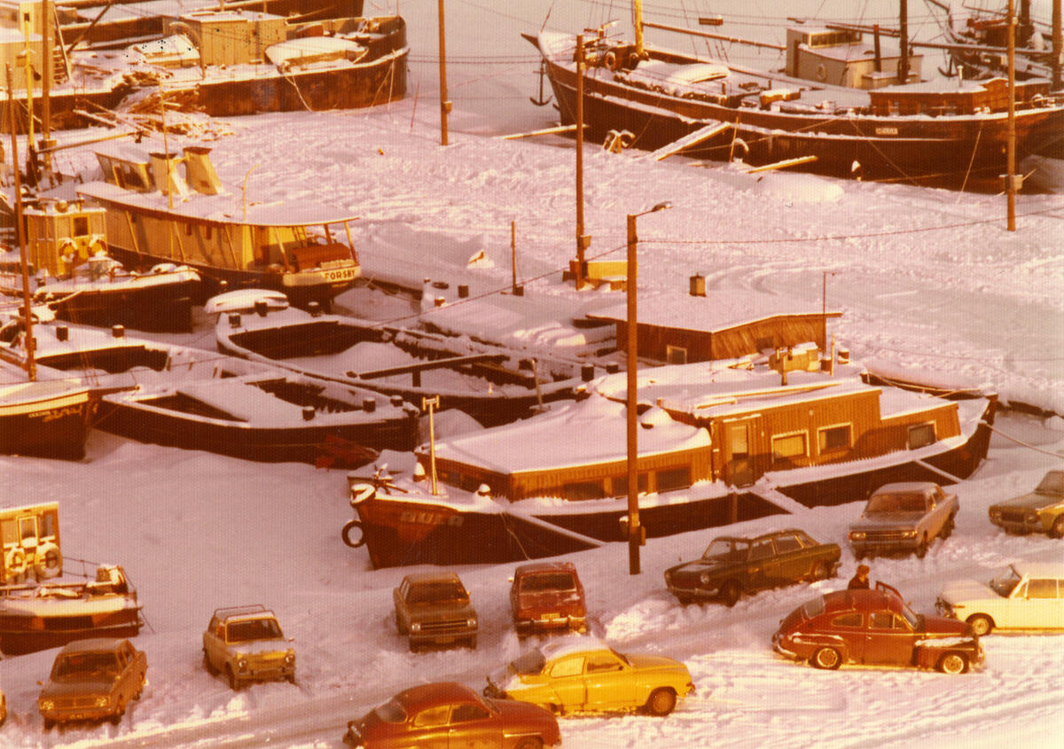 Several houseboats moored very close to each other. There are cars on the seashore. The ground is covered with snow.