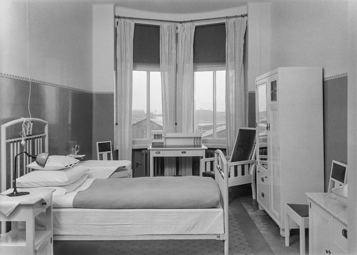 Yhden hengen potilashuone, jossa valkoinen sänky, yöpöytä, vaatekaappi ja pesukomuutti. Ikkunaerkkerin edessä on siro valkoinen kirjoituspöytä. Huoneessa on kolme tuolia ja lattialla matto.
