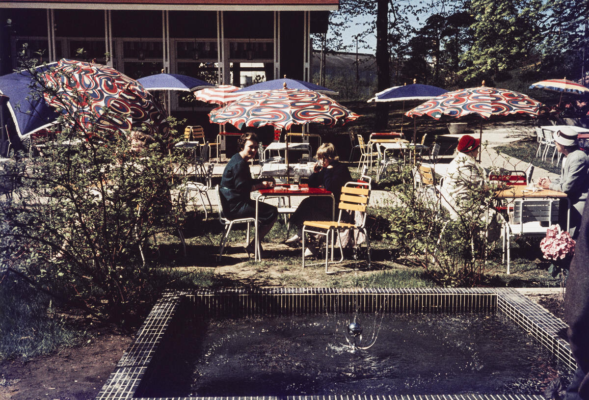 Kafé Tölöstrands gård och springbrunn. Kafékunder sitter vid borden under parasoll och njuter förfriskningar.