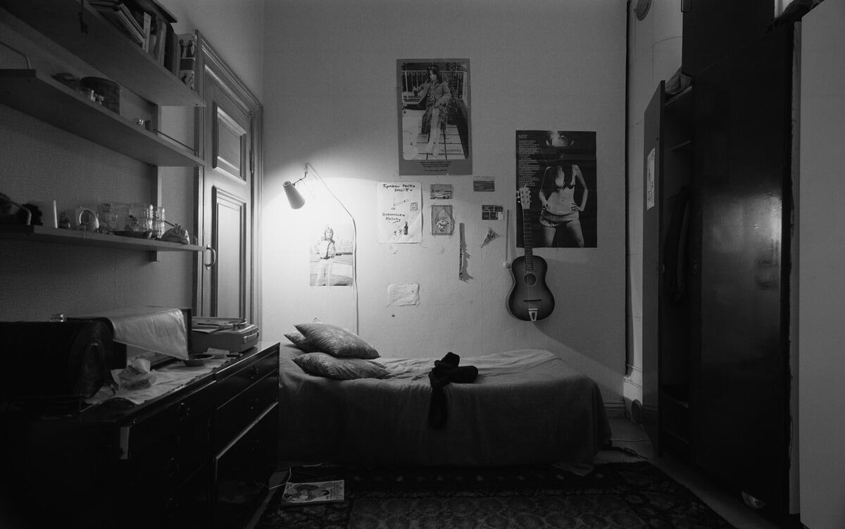 A teenager’s room with a bed, a table and a set of shelves. There are posters and a guitar on the wall.