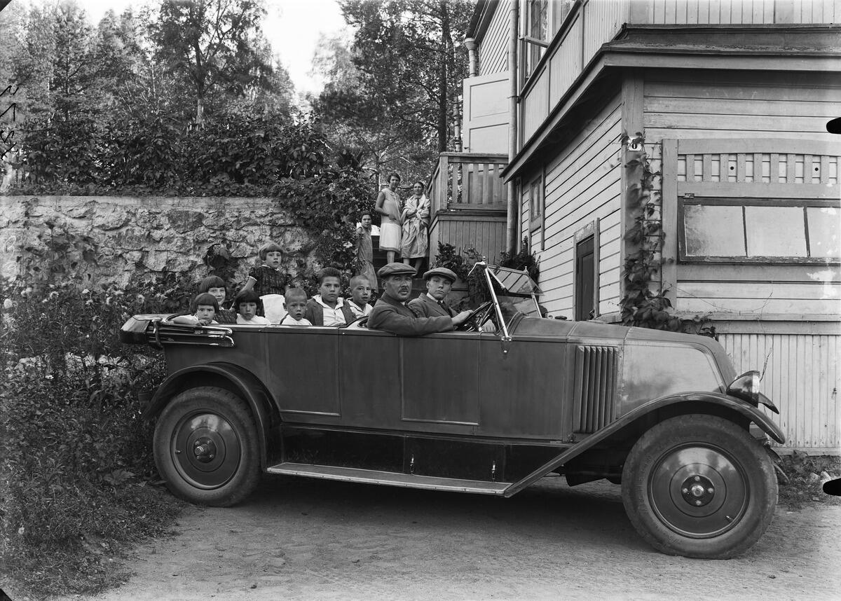 En öppen bil med två baksäten framför hörnet av en trävilla. Det sitter åtta personer i bilen, två kvinnor står på trappan i bakgrunden.