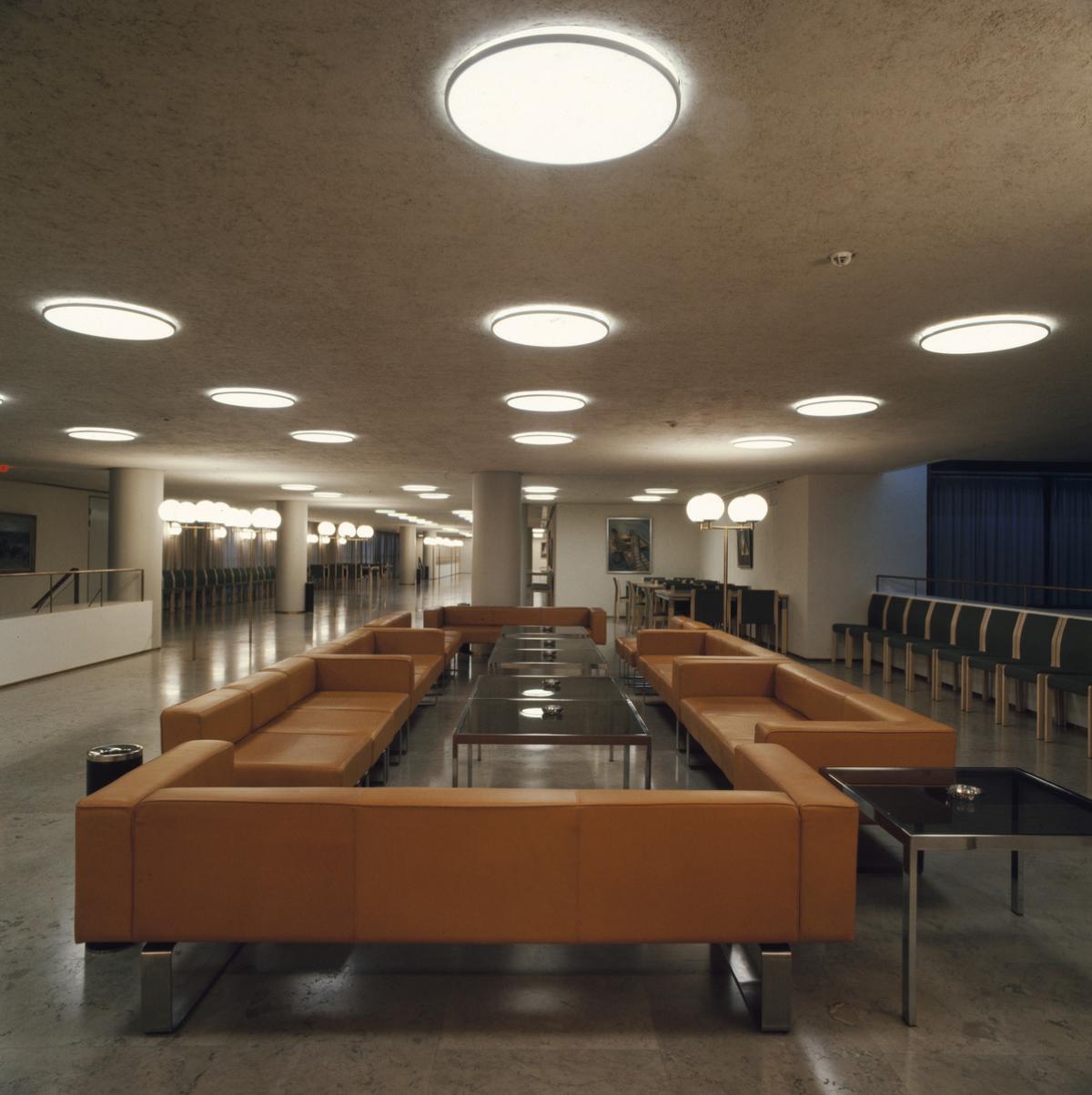 Kuva Helsingin kaupungintalon käytävältä. Tilaa hallitsee suuri, tummanruskea sohvaryhmä. Käytävän kattoon on upotettu pyöreitä valaisimia.