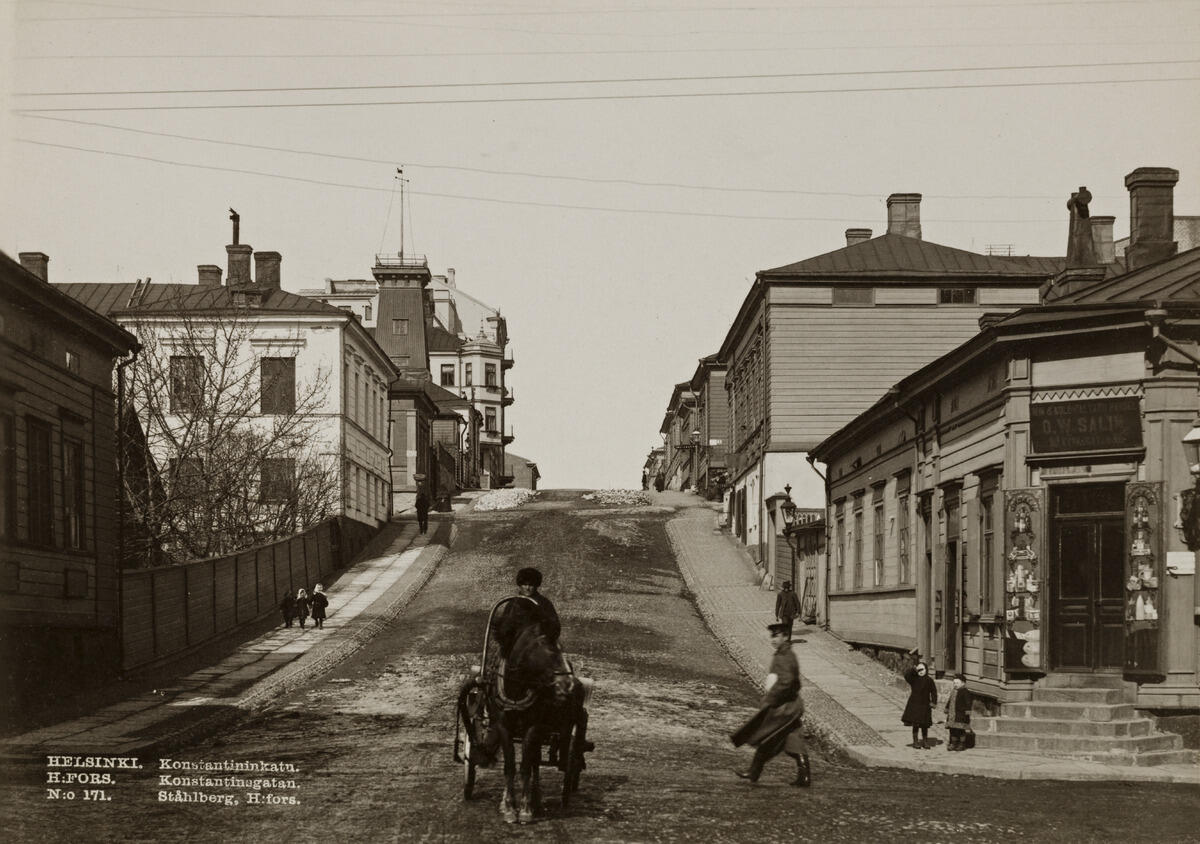 Konstantininkatu (now Meritullinkatu) 5 and 7 (on right) and 10, 12 and 14 (on left). The Kruununhaka fire tower (now Meritullinkatu 12) can be seen on the left side of the street. Kirkkokatu is at the foreground, and the Win & Kolonialvaruhandel O.W. Salin shop at the corner to the right.