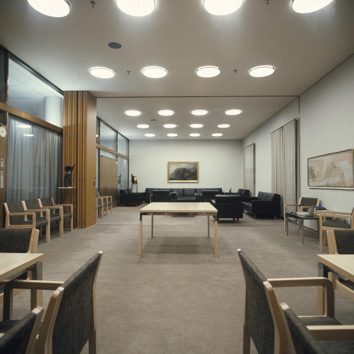 Sisäkuva Helsingin kaupungintalon toimistotiloista. Vaalean huoneen katossa on pyöreitä, kattoon upotettuja valaisimia. Tilassa on pöytiä ja tumma sohvaryhmä sekä lasiseinä käytävälle.