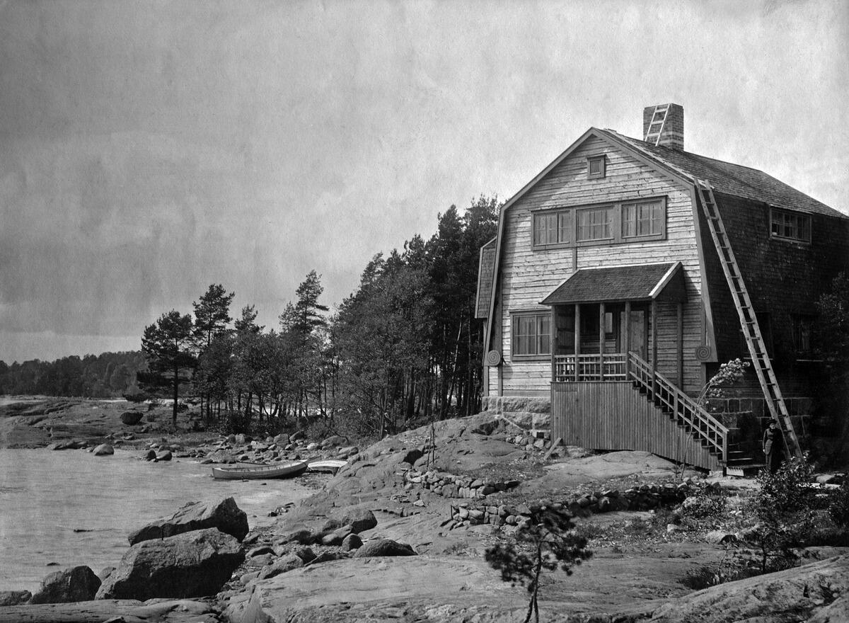 En tvåvåningsträvilla på klippor vid stranden. Till vänster syns havet och träd. En kvinna står på trappan. 