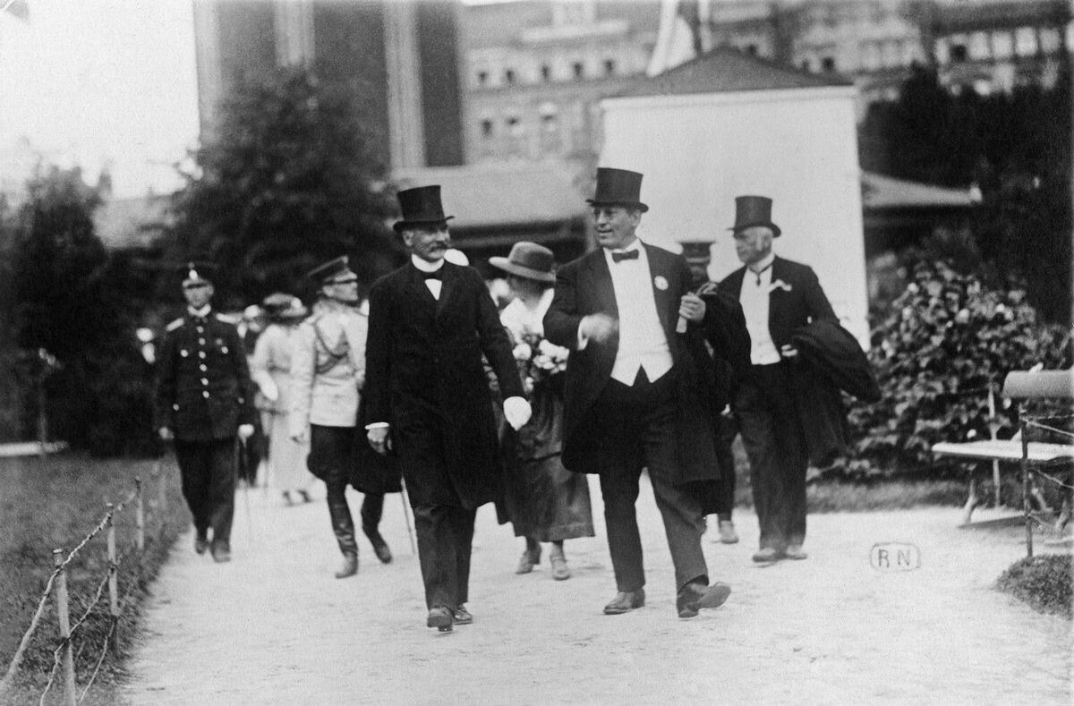 I förgrunden spatserar män klädda i cylinderhattar och festliga kläder. Bakom dem uniformklädda män och festklädda kvinnor.