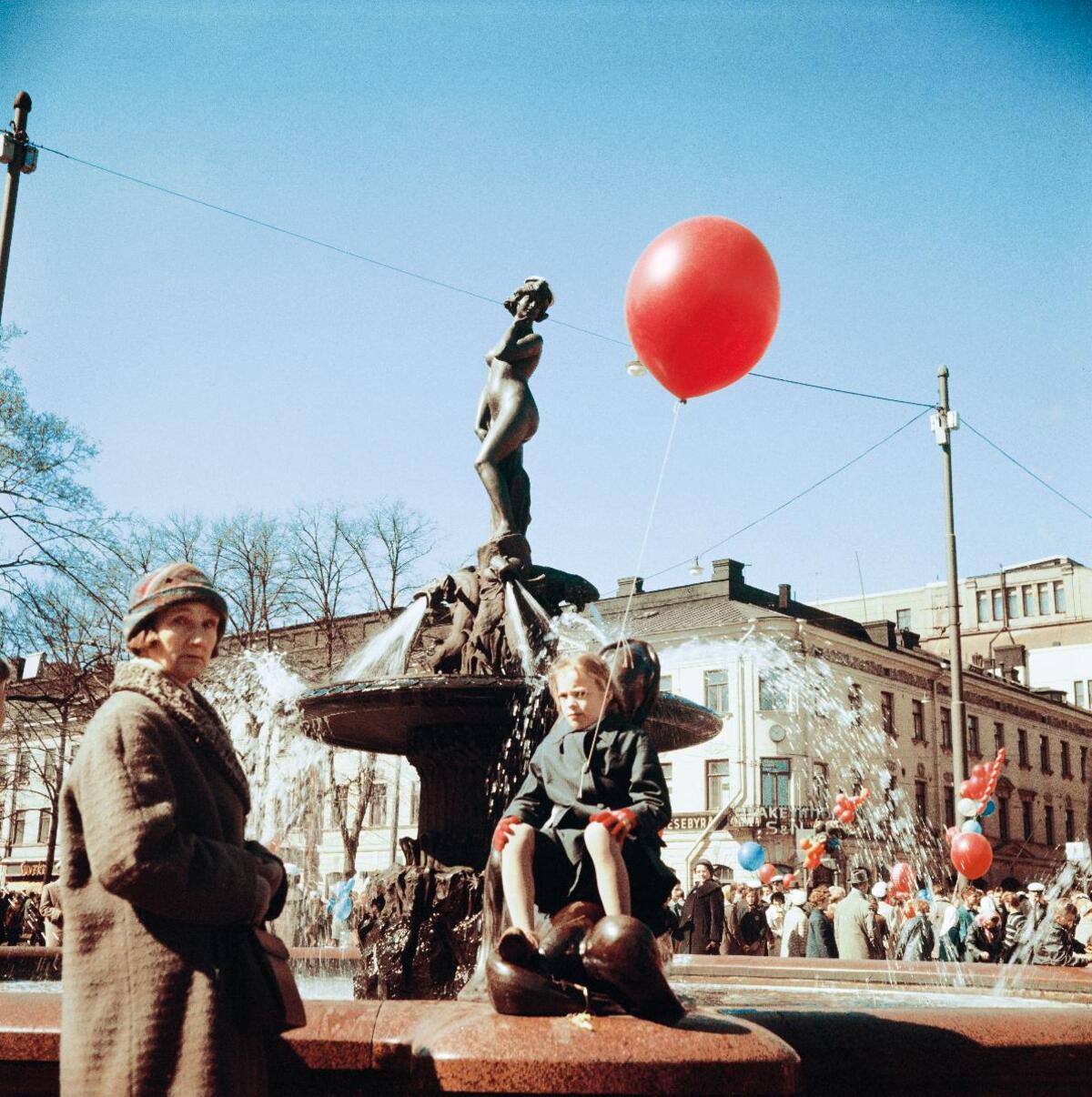A girl with a red balloon sitting on a seal statue at a fountain. A woman standing to the left in the front of the picture.