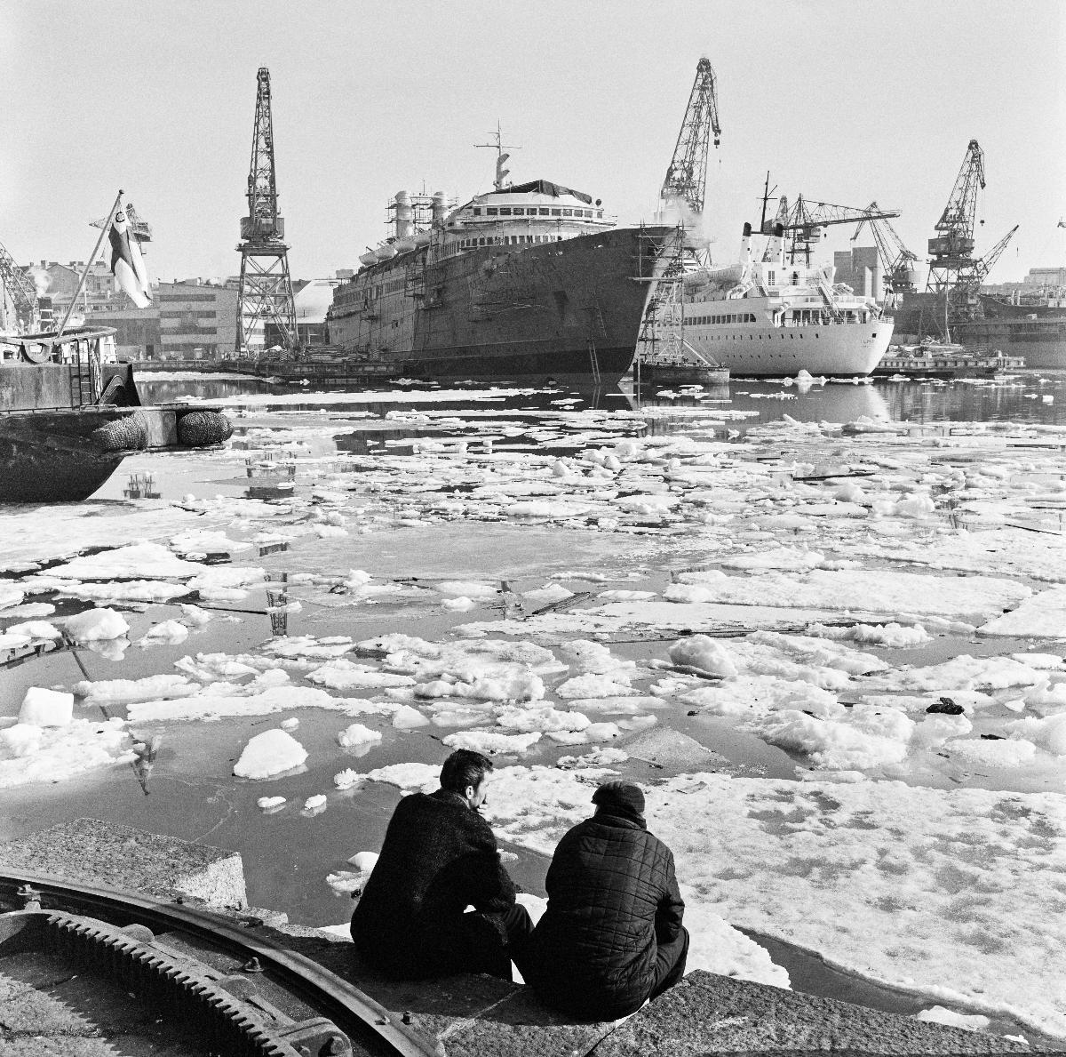 Två män sitter vid Sandvikskajen. I bakgrunden syns Wärtsiläs varv i Sandviken. Det vita fartyget är m/s Ilmatar. Foto: Helsingfors stadsmuseum / Volker von Bonin