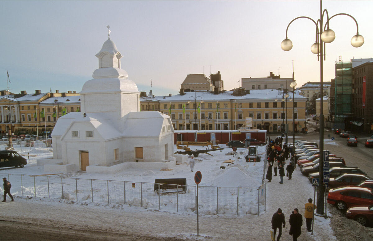 Näkymä talviselle Senaatintorille. Sen läntiselle puoliskolle on rakennettu lumesta kopio Ulrika Eleonoran kirkosta, joka aikanaan sijaitsi samalla paikalla. Luminen kirkkomaa on aidattu, koska rakentaminen on vielä hieman kesken. Torin etelälaidalla osin kirkon takana on työmaaparakkeja. Aitauksen ulkopuolella ihmiset ovat pysähtyneet katsomaan näkyä. Torin laitaan on pysäköity yksityisautoja. Ilta-aurinko paistaa viistosti valaisten kirkon ylempiä osia.