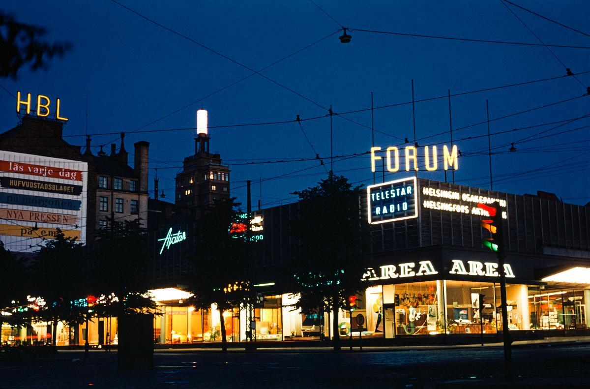 Lasipalatsin rakennus Mannerheimintiellä iltavalaistuksessa. Rakennuksen kulmassa matkatoimisto Area. Taustalla näkyy kyltti Forum, Telestar Radio, Ajatar ja vasemmalla Hufvudstadsbladetin kyltti HBL.