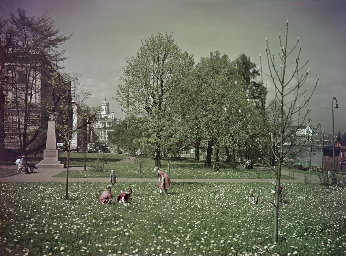 Leikkiviä lapsia Tähtitorninmäen puistossa. Kuvan reunassa muistopaasi, jonka päällä rautaristi.