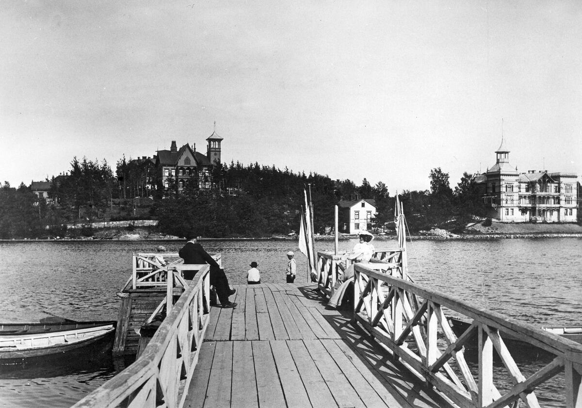 Töölönlahden rannassa puinen venelaituri, jonka reunoilla olevilla penkeillä istuu mies ja nainen. Laiturin päässä seisoo kolme lasta. Vastarannalla kaksi Eläintarhan huvilaa.