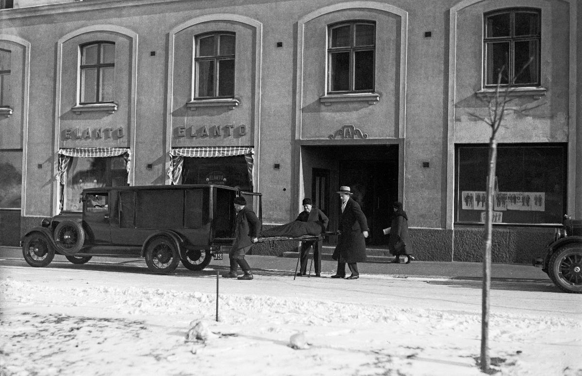 Luminen katu ja jalkakäytävä, jolle on pysähtynyt ambulanssi tai ruumiinkuljetusauto. Kolme miestä nostaa paareilla makaavaa, tummalla huovalla peitettyä ruumista auton takaosaan. Taustalla kävelee ohikulkija.