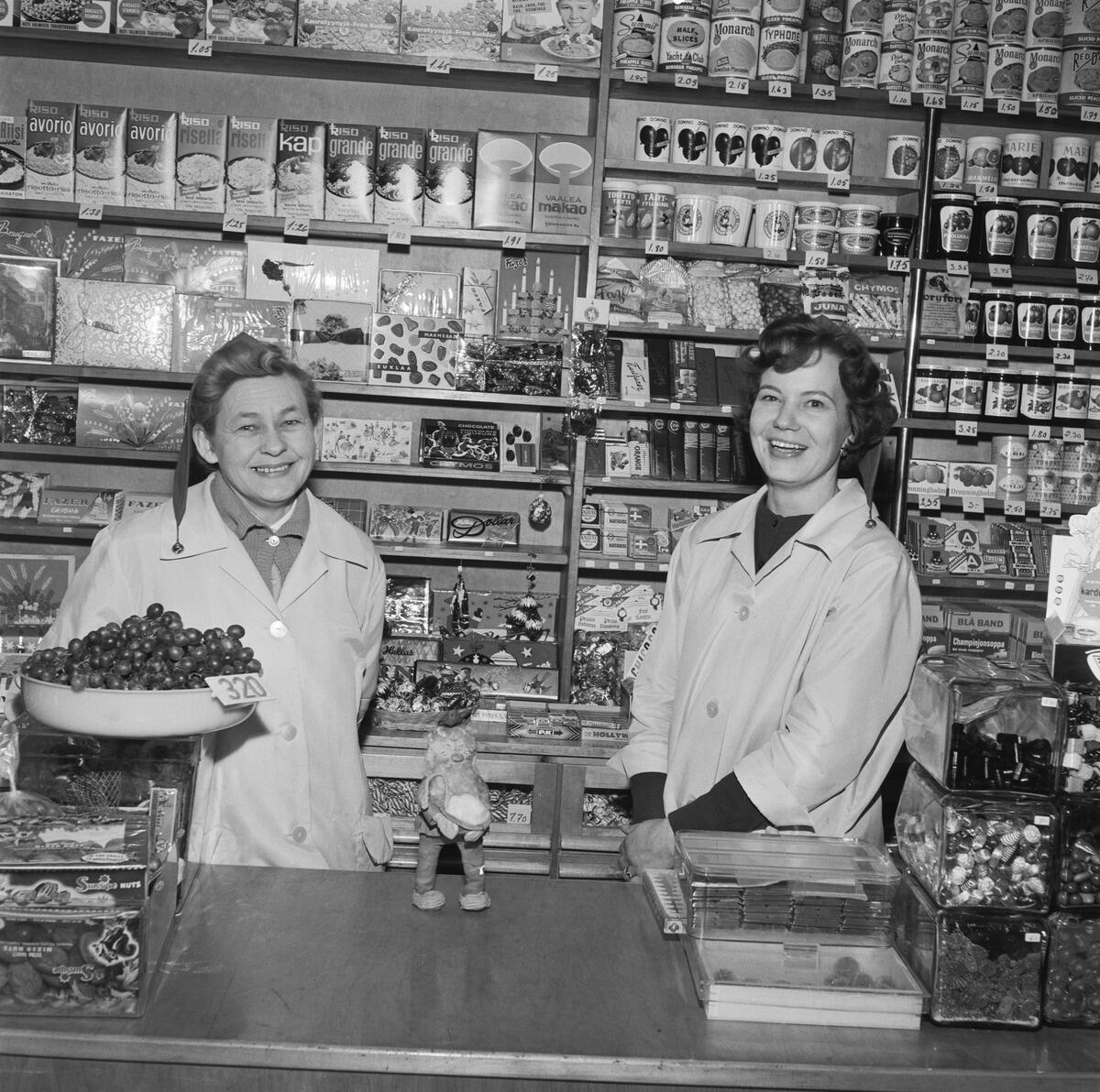Kaksi hymyilevää naista seisoo kaupan tiskin takana. Toisella on tonttulakki päässään ja viinirypälevati kädessään. Naisten takana on hyllyjä täynnä ruokapakkauksia ja konvehtirasioita.