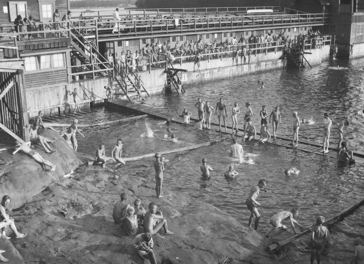 Paljon poikia ja miehiä uimassa kesäisenä päivänä puurakenteisessa uimalassa.