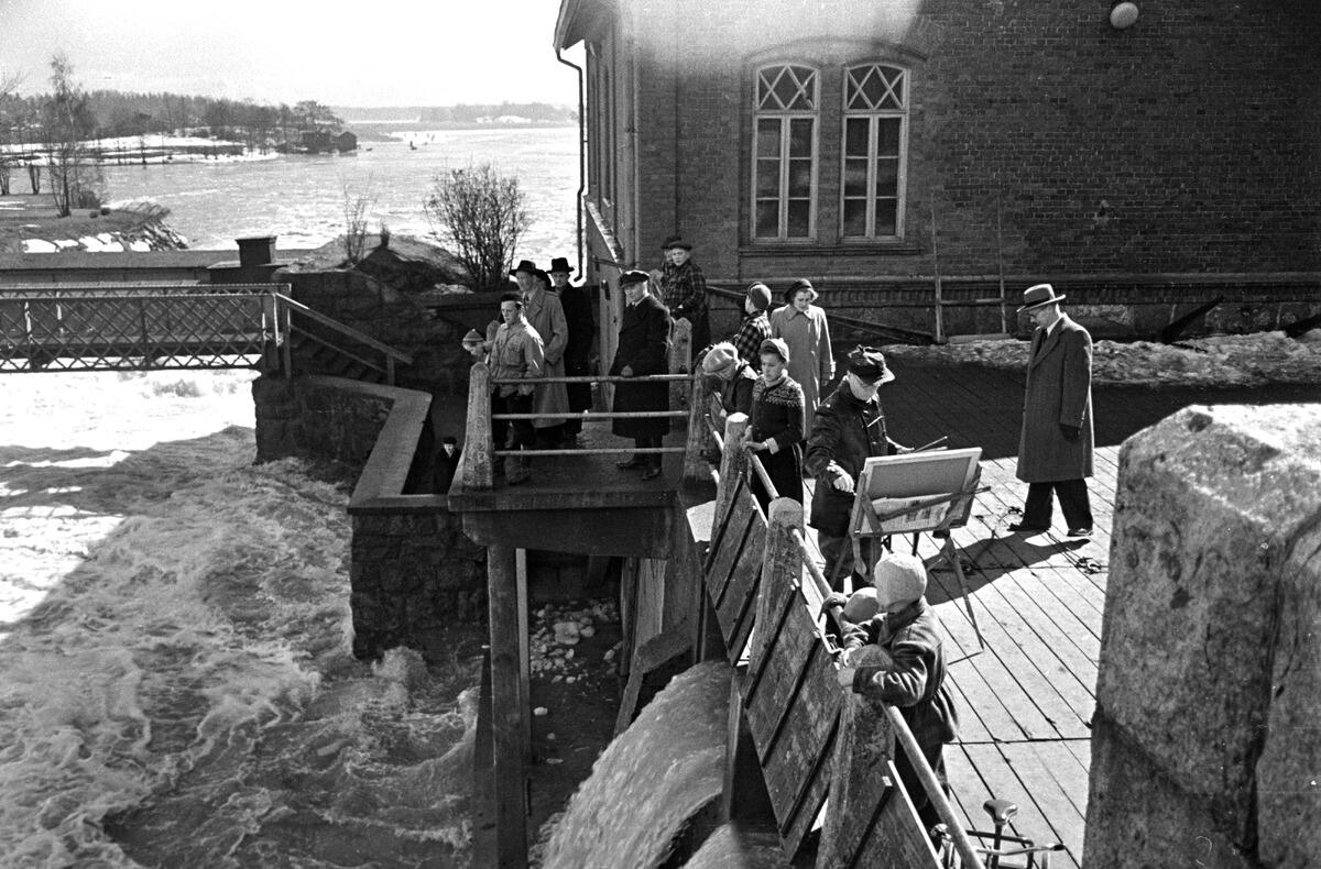 Ihmisiä kokoontuneena kuohuvan Vanhankaupunginkosken äärelle jäidenlähdön aikaan. Yksi mies seisoo maalaustelineen ääressä maalaamassa.