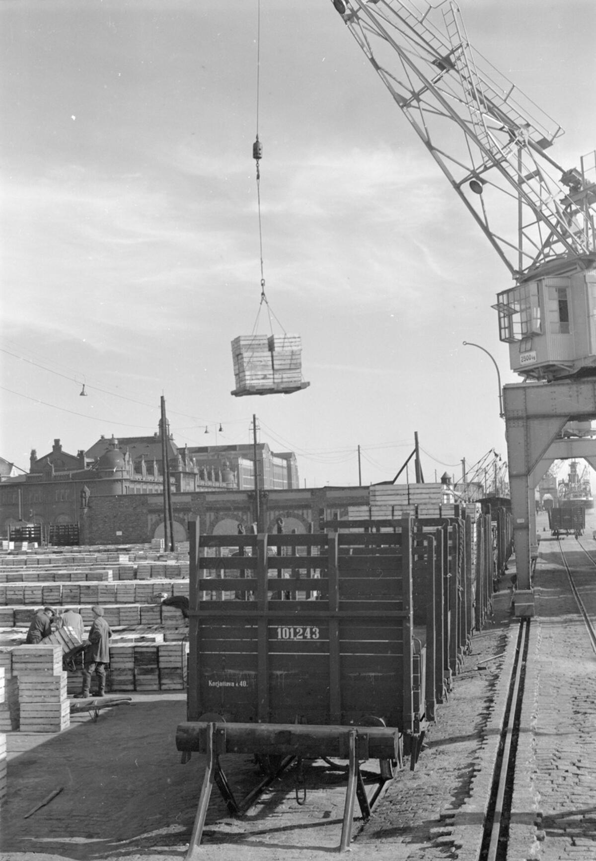 Till vänster syns bredstaplar och hamnarbetare. I förgrunden står en öppen godsvagn som håller på att lastas med hjälp av en lyftkran.